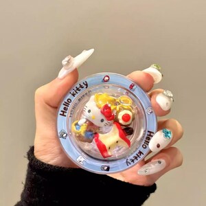 May include: A clear, round case with a blue rim featuring a Hello Kitty design. The case contains miniature Hello Kitty figures and food items. The text "Hello Kitty" is printed on the rim. The text "YQQXCase" is printed at the bottom.