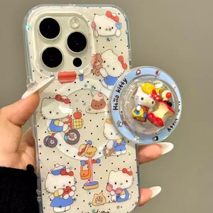 May include: Clear phone case with a Hello Kitty design. The case features multiple illustrations of Hello Kitty in various poses, with a matching pop socket. The pop socket has a blue border and a 3D Hello Kitty design. The text "Hello Kitty" is visible.