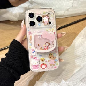 以下が含まれることがあります： ピンクのハローキティのデザインの透明な携帯電話ケース。ケースにはポップアップグリップと、クマ、カップケーキ、リンゴなど、さまざまなハローキティのキャラクターイラストが描かれています。「Cute」という文字も表示されています。