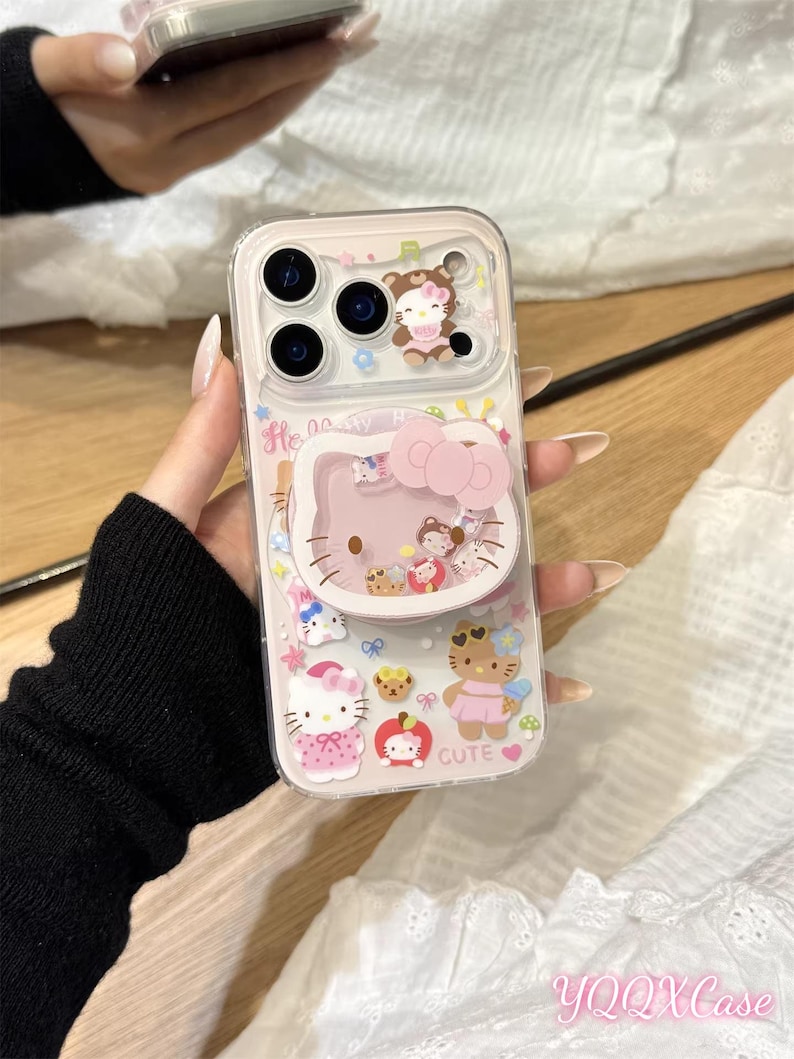 以下が含まれることがあります： ピンクのハローキティのデザインの透明な携帯電話ケース。ケースにはポップアップグリップと、クマ、カップケーキ、リンゴなど、さまざまなハローキティのキャラクターイラストが描かれています。「Cute」という文字も表示されています。
