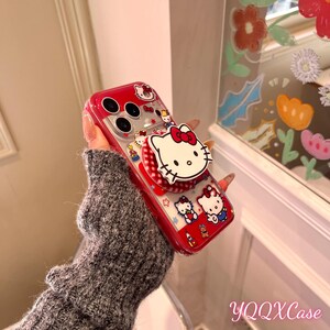 May include: A red and clear phone case with Hello Kitty designs. The case has a pop-up grip with a white Hello Kitty face and red bow. Additional Hello Kitty characters and stars decorate the case. The text "YQQXCase" is visible.