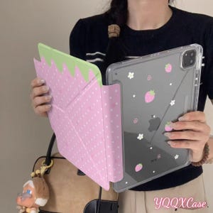 May include: A pink and green strawberry-themed iPad case and a clear iPad case with pink strawberry and white flower designs. The strawberry case is open, revealing a dotted pattern. The clear case has the text "YQQXCase".