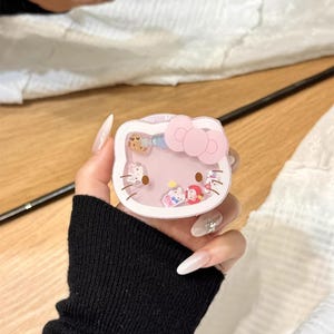 以下が含まれることがあります： ピンクのリボンが付いた、透明、ピンク、白のハローキティ型の携帯電話ケース。ケースには、小さなカラフルなキャラクターフィギュアが入っています。「YQQXCase」の文字が右下に表示されています。