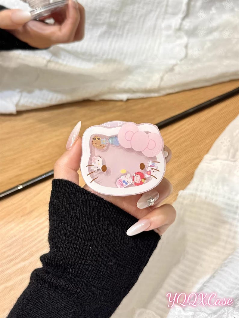 以下が含まれることがあります： ピンクのリボンが付いた、透明、ピンク、白のハローキティ型の携帯電話ケース。ケースには、小さなカラフルなキャラクターフィギュアが入っています。「YQQXCase」の文字が右下に表示されています。