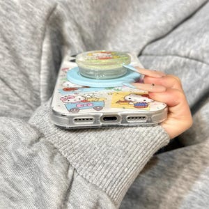 Puede incluir: Una funda transparente para tel&eacute;fono con un dise&ntilde;o de dibujos animados y un soporte para tel&eacute;fono de color azul claro. La funda presenta un patr&oacute;n colorido con personajes de dibujos animados. El soporte tiene una parte superior transparente con una imagen de dibujos animados.