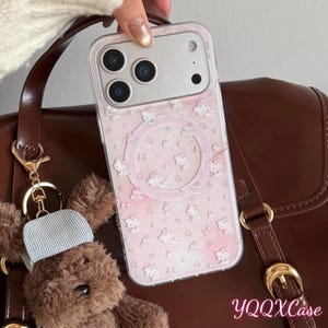 Puede incluir: Una funda transparente para tel&eacute;fono con fondo rosa y un estampado de personajes de dibujos animados blancos y pasteles. La funda est&aacute; sujeta por una mano y la c&aacute;mara del tel&eacute;fono es visible. La funda est&aacute; junto a un bolso marr&oacute;n y un llavero de oso de peluche.