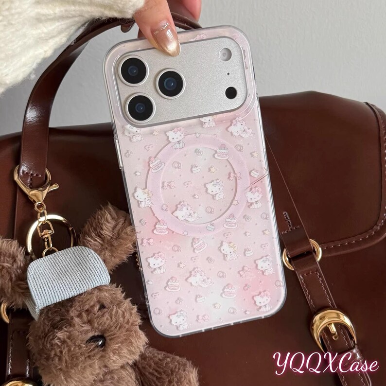 Puede incluir: Una funda transparente para tel&eacute;fono con fondo rosa y un estampado de personajes de dibujos animados blancos y pasteles. La funda est&aacute; sujeta por una mano y la c&aacute;mara del tel&eacute;fono es visible. La funda est&aacute; junto a un bolso marr&oacute;n y un llavero de oso de peluche.