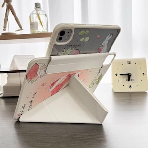 May include: A white phone case with a strawberry and bunny design, propped up on a white stand. The phone case has a light gray back with a bunny and cherry design. The stand is folded into a triangular shape.