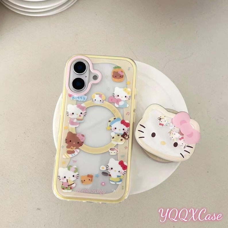 May include: A clear phone case with a yellow border and a pop-up grip featuring Hello Kitty characters. The case has multiple Hello Kitty designs. The pop-up grip is white with a pink bow and Hello Kitty face.