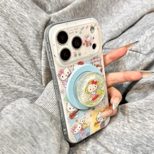 Puede incluir: Funda transparente para tel&eacute;fono con un dise&ntilde;o de Hello Kitty y un soporte emergente azul claro. La funda tiene un borde blanco y un patr&oacute;n repetido de personajes de Hello Kitty, flores y otros elementos decorativos.