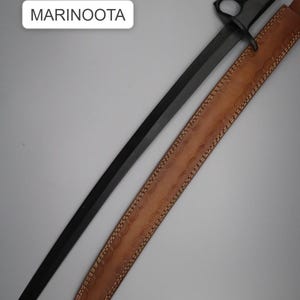 Puede incluir: Una daga negra con un mango de nudillos y una funda de cuero marrón. La hoja es larga y recta. La funda es de color canela con un patrón decorativo. El texto "MARINOOTA" está en un rectángulo blanco.