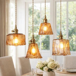 May include: Four pendant lights with amber glass shades hang above a dining table. The shades have textured surfaces and are illuminated, casting a warm glow. The lights are suspended by brown cords and brass fixtures.