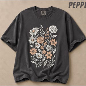 Puede incluir: Una camiseta de manga corta gris carbón con un estampado gráfico central de varias flores silvestres en tonos blancos y melocotón suave. La camiseta tiene cuello redondo y una etiqueta en el cuello interior que dice Comfort Colors y S-P-CH.