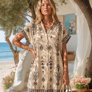 May include: Beige button-down shirt dress with black ornamental patterns. The short-sleeved dress features a collared neckline and a button-down front. A woven tote bag is held in the hand. The background includes a beach scene with ocean and trees.