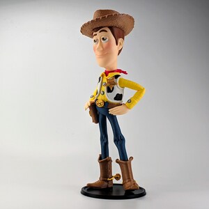 May include: A standing figurine of a cowboy character wearing a brown hat, a yellow long-sleeved shirt, a white vest with black cow-print spots, a red neckerchief, blue pants, and brown cowboy boots with gold spurs, mounted on a black circular base.