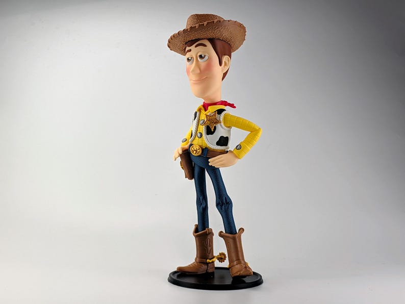 May include: A standing figurine of a cowboy character wearing a brown hat, a yellow long-sleeved shirt, a white vest with black cow-print spots, a red neckerchief, blue pants, and brown cowboy boots with gold spurs, mounted on a black circular base.