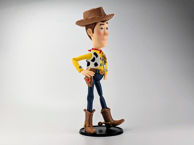 May include: A plastic figurine of a cowboy character wearing a brown textured hat, a yellow long-sleeve shirt, a white vest with black cow-print spots, a red neckerchief, a gold sheriff badge, a brown belt with a gold buckle, and blue pants.