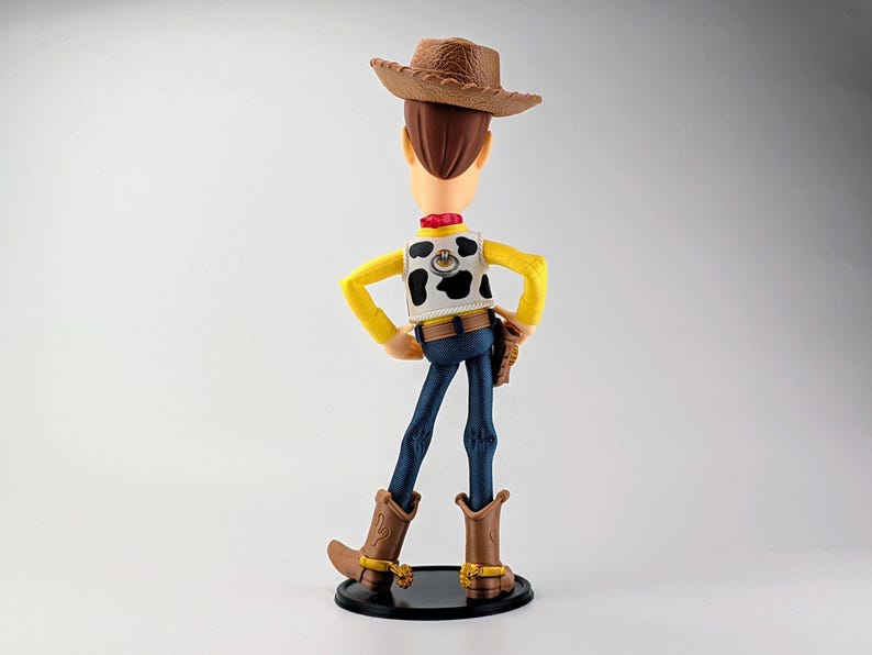 May include: A cowboy action figure seen from behind, wearing a brown textured hat, a yellow long-sleeved shirt, a white vest with black cow-print spots, blue pants, and brown boots with gold spurs, standing on a circular black base.