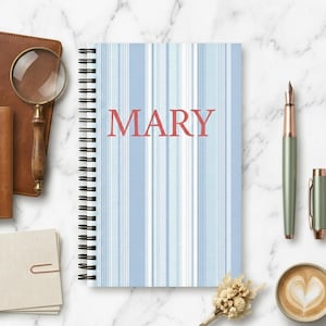 Pode incluir: Um caderno de espiral com um padrão de riscas verticais em azul claro e branco, com o nome MARY impresso em letras maiúsculas vermelhas no centro. O caderno está posicionado entre vários artigos de escritório numa superfície de mármore.