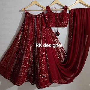 Puede incluir: Un conjunto burdeos de tres piezas, que incluye una falda, un top corto y una bufanda drapeada, todo colgado de perchas de madera. La falda y el top están adornados con detalles dorados. El texto "RK designer" es visible.