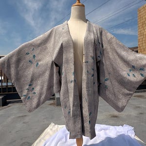May include: A gray kimono-style robe with a subtle pattern and teal leaf-like accents. The robe is displayed on a wooden mannequin, with its sleeves spread wide. The background shows a blue sky and a building.