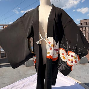 May include: Black kimono with floral and leaf patterns in white, orange, and yellow. The kimono is displayed on a mannequin against a blue sky background. The sleeves are wide and the garment is open in the front.