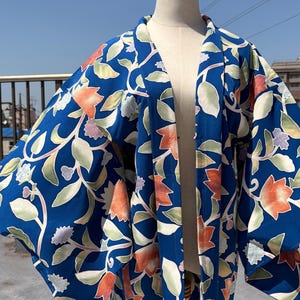 May include: A blue kimono-style jacket with a floral pattern in orange, green, and white. The jacket is displayed on a mannequin against a clear blue sky. The floral design is detailed and covers the entire garment.