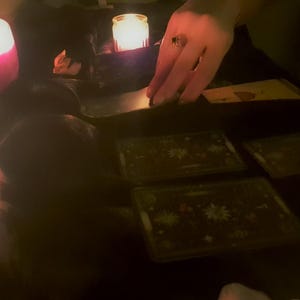 May include: A set of rectangular tarot cards with dark backgrounds and intricate star patterns, arranged on a surface illuminated by the warm, glowing light of several small candles, creating a dim and focused atmosphere for card reading.