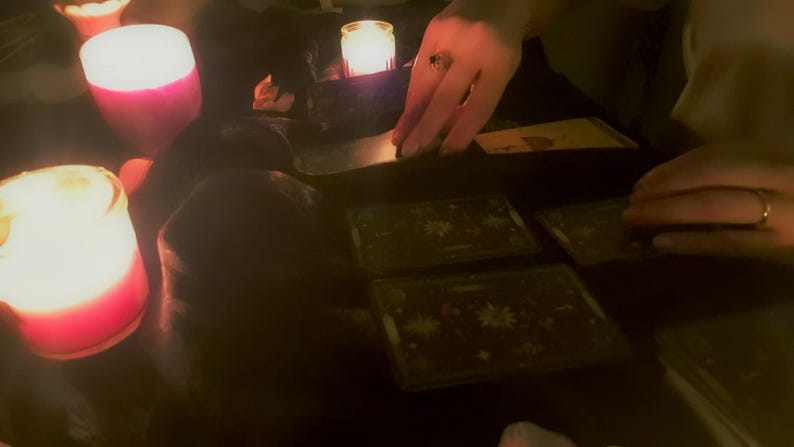 May include: A set of rectangular tarot cards with dark backgrounds and intricate star patterns, arranged on a surface illuminated by the warm, glowing light of several small candles, creating a dim and focused atmosphere for card reading.