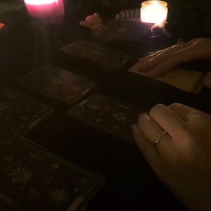 May include: A set of rectangular tarot cards with dark backgrounds and intricate white celestial illustrations, including stars and moons, arranged on a surface illuminated by the warm glow of two lit candles in glass containers.