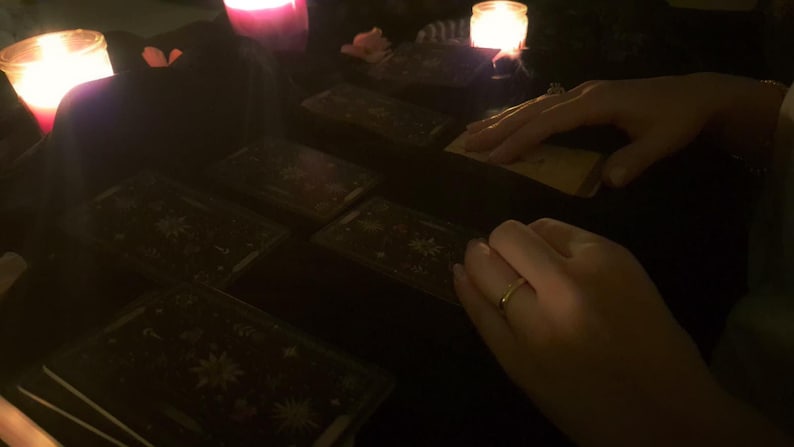 May include: A set of rectangular tarot cards with dark backgrounds and intricate white celestial illustrations, including stars and moons, arranged on a surface illuminated by the warm glow of two lit candles in glass containers.
