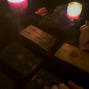 May include: A set of rectangular tarot cards arranged on a dark surface, including a card featuring two swords and another displaying a single chalice. The scene is illuminated by the warm, soft glow of several lit candles, creating a dim atmosphere.