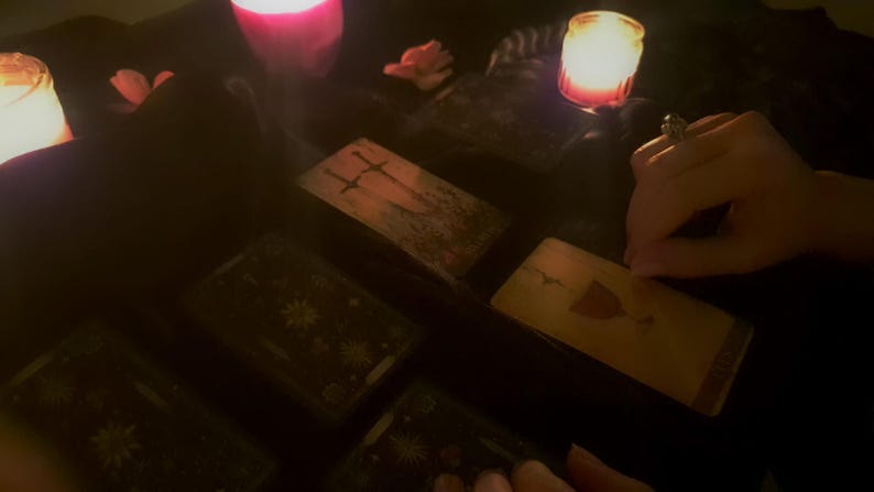 May include: A set of rectangular tarot cards arranged on a dark surface, including a card featuring two swords and another displaying a single chalice. The scene is illuminated by the warm, soft glow of several lit candles, creating a dim atmosphere.
