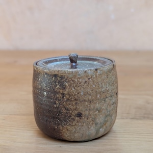 Puede incluir: Un pequeño tarro de cerámica hecho a mano con tapa. El tarro es una mezcla moteada de marrón y beige, con una banda marrón más oscura cerca de la base. La tapa tiene un pequeño pomo y coincide con la combinación de colores del tarro.