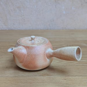 Peut inclure: Une théière à poignée latérale fabriquée en argile tachetée avec une finition naturelle couleur terre cuite et crème. La théière présente un corps rond, un bec court, un couvercle plat avec un petit bouton et une large poignée cylindrique.