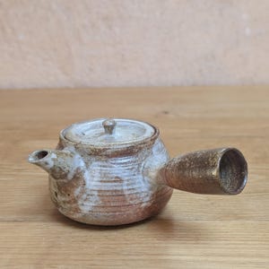 Może przedstawiać: Ceramiczny czajniczek z bocznym uchwytem, pokryty nakrapianą brązowo-białą glazurą. Czajniczek ma okrągły korpus, mały dzióbek oraz dopasowaną pokrywkę z centralnym uchwytem. Powierzchnia posiada widoczne poziome żłobienia i teksturę.