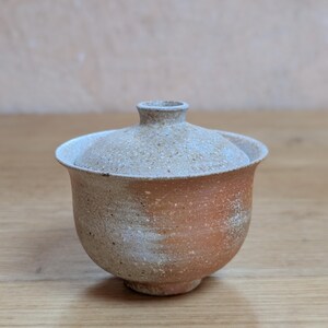Könnte beinhalten: Eine kleine, runde Keramik-Teeschale mit passendem Deckel, die eine texturierte, gesprenkelte Oberfläche mit einem Farbverlauf aus erdigem Terrakotta-Orange und gedämpften Beigetönen aufweist. Die Schale hat einen ausgestellten Rand.