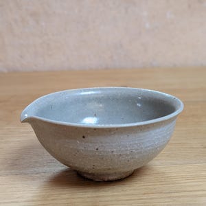 Può includere: Una piccola ciotola in ceramica rotonda con smalto grigio chiaro maculato e interno liscio. L'esterno presenta una finitura ruvida e terrosa con macchie scure visibili. Un piccolo beccuccio integrato si trova sul bordo sinistro.