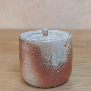 May include: A handmade ceramic jar with a lid. The jar is cylindrical with a rounded base and a small knob on the lid. The colors are a blend of beige, brown, and reddish-brown, with a speckled texture.