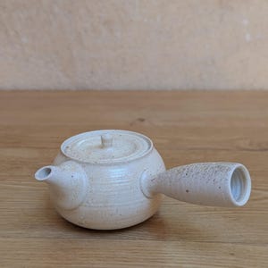 Op de afbeelding: Een theepot met zijhandvat gemaakt van gebroken wit keramiek met een gespikkelde textuur. De theepot heeft een rond lichaam, een kleine tuit, een bijpassend rond deksel met een centrale knop en een dik, taps toelopend handvat aan de zijkant.