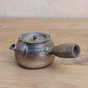 Può includere: Una piccola teiera in ceramica marrone con un manico laterale e un coperchio. La teiera ha un beccuccio e un aspetto rustico e strutturato. Il manico è in legno. La teiera è appoggiata su una superficie di legno.