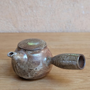 Może przedstawiać: Okrągły, nakrapiany brązowy czajniczek ceramiczny z bocznym uchwytem i pasującą pokrywką. Powierzchnia posiada nakrapiane, ziemiste szkliwo z różnicami w odcieniach, w tym ciemnobrązowe plamki i jaśniejsze beżowe obszary na korpusie.
