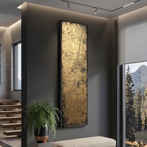 May include: A rectangular abstract artwork with a gold and black textured design, framed in black. The piece is mounted on a dark grey wall. A fern in a black and copper pot is in the foreground.