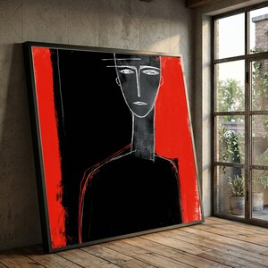 May include: Abstract art print featuring a minimalist portrait in black and white with a red vertical stripe. The artwork is framed in a black frame and leans against a textured wall.
