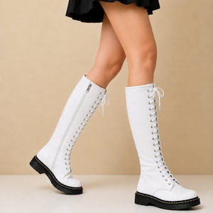 May include: A pair of knee-high white boots featuring a lace-up front with silver eyelets, white laces, and a side zipper closure. The boots have a thick black lug sole with yellow stitching along the welt, providing a sturdy base for the footwear.