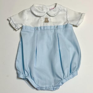 Könnte beinhalten: Ein Baby-Strampler in Weiß und Hellblau. Das Oberteil ist weiß mit einem Bubikragen und einem kleinen gestickten Teddybären. Der Unterteil ist hellblau mit Falten und einer Ballonform. Der Strampler hat kurze Ärmel und Knöpfe.