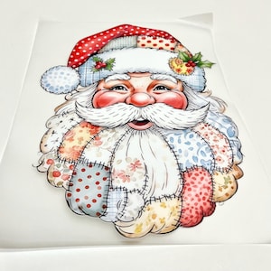 Pode incluir: Uma transferência decorativa apresentando um rosto de Pai Natal com um design de patchwork. A imagem exibe um gorro vermelho com bolinhas brancas e uma barba composta por vários segmentos de tecido floral e estampado em tons pastel.