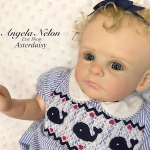 Puede incluir: Una muñeca bebé realista con ojos azules y cabello rubio rizado, vestida con un vestido a rayas azul y blanco con un diseño de ballena. La muñeca tiene un lazo azul en el pelo. El texto en la imagen dice "Angela Nelon Etsy Shop Asterdaisy."