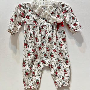 Può includere: Pagliaccetto bianco per neonati con un motivo floreale di rose rosse e foglie verdi. Il pagliaccetto presenta maniche lunghe, polsini arricciati e un colletto rifinito in pizzo. Un piccolo fiocco rosso adorna il davanti.