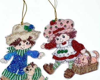 May include: Two glittery Christmas ornaments featuring cartoon characters. One is a boy in blue overalls with a dog, the other is a girl in a strawberry-themed outfit with a cat in a basket. Both have gold hanging strings.